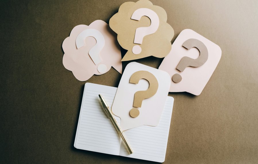 Auf einem Tisch liegt ein aufgeschlagenes Schulheft. Darüber sind übergroße Sprechblasen abgebildet, die Fragezeichen enthalten.